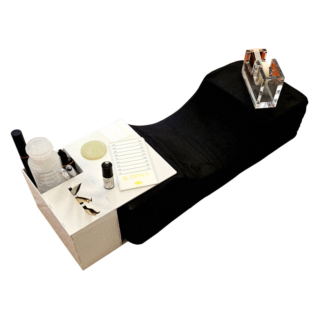 Angled view of white acrylic eyelash extension pillow stand with tray and organizer compartments holding lash tools and products